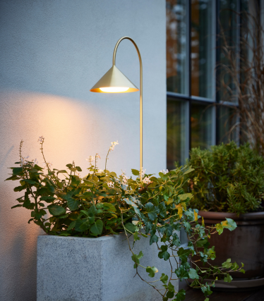 Leuchtende Outdoor-Lampe von Frandsen neben einem bepflanzten Betonkübel auf einer Terrasse vor moderner Hausfassade.