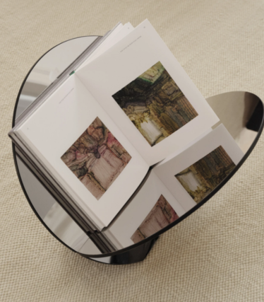 Runder Beistelltisch aus dunklem Glas mit aufgeschlagenem Kunstbuch, auf beigem Teppich.